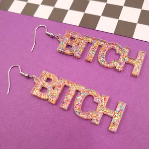 May include: A pair of dangle earrings with the word "BITCH" spelled out in clear plastic with colourful glitter embedded in the letters. The earrings have silver hooks.