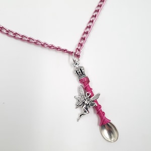 Puede incluir: Un collar de cadena rosa con un dije de cuchara de plata envuelto en alambre rosa. La cuchara tiene una corona y un dije de hada.