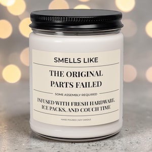Könnte beinhalten: Eine weiße Sojakerze in einem klaren Glasgefäß mit schwarzem Deckel. Das Etikett trägt die Aufschrift: SMELLS LIKE THE ORIGINAL PARTS FAILED, SOME ASSEMBLY REQUIRED, INFUSED WITH FRESH HARDWARE, ICE PACKS, AND COUCH TIME, HAND POURED | SOY CANDLE.