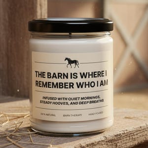 Op de afbeelding: Een doorzichtige glazen kaarsenpot met een zwarte deksel en witte was. Het etiket bevat een paardenicoon en de tekst: THE BARN IS WHERE I REMEMBER WHO I AM, INFUSED WITH QUIET MORNINGS, STEADY HOOVES, AND DEEP BREATHS, 219% NATURAL, BARN THERAPY, HAND-POURED.