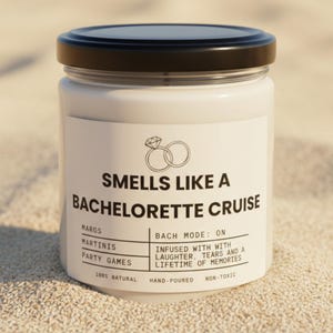 May include: A white candle in a clear glass jar with a black lid. The label features a diamond ring icon and the text: SMELLS LIKE A BACHELORETTE CRUISE, MARGS, MARTINIS, PARTY GAMES, BACH MODE: ON, INFUSED WITH LAUGHTER, TEARS AND A LIFETIME OF MEMORIES.