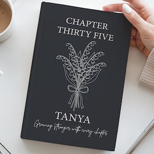 May include: A black journal with white text and a floral illustration. The cover reads "CHAPTER THIRTY FIVE" above a bouquet of flowers and the name "TANYA" with the phrase "Growing stronger with every chapter."