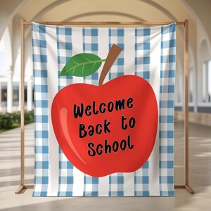 May include: A fabric backdrop featuring a blue and white gingham pattern with a large red apple graphic in the centre. The apple has a brown stem and a green leaf, with the black text Welcome Back to School printed across the front of the apple.