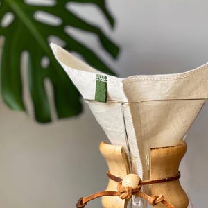 May include: A clear glass coffee maker featuring a reusable off-white fabric filter with a small green tab. The neck of the glass vessel is wrapped in a light-colored wooden collar secured by a brown leather cord tied with a wooden bead.