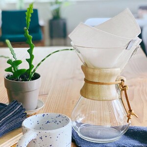 May include: A glass pour-over coffee maker with a light wood collar and a leather tie, featuring a white fabric filter inside. Beside it sits a white ceramic mug with blue speckles, a small potted green plant, and a stack of dark blue fabric.