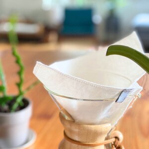 May include: A reusable off-white fabric coffee filter with stitched edges and a small grey tab, positioned inside a clear glass pour-over coffee maker with a wooden collar and a leather tie, while a green spout pours water into the filter.