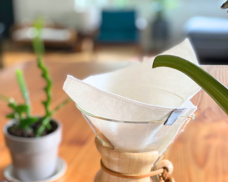 May include: A reusable off-white fabric coffee filter with stitched edges and a small grey tab, positioned inside a clear glass pour-over coffee maker with a wooden collar and a leather tie, while a green spout pours water into the filter.