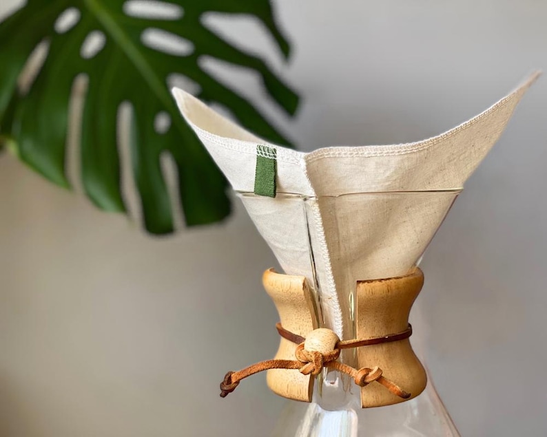 May include: A clear glass coffee maker featuring a reusable off-white fabric filter with a small green tab. The neck of the glass vessel is wrapped in a light-colored wooden collar secured by a brown leather cord tied with a wooden bead.