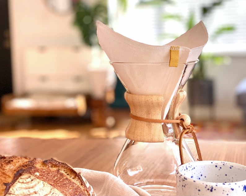 May include: A clear glass pour-over coffee maker with a light-colored wooden collar secured by a tan leather cord. A white fabric coffee filter is inserted into the top, featuring a small yellow fabric tab on the side rim of the filter.