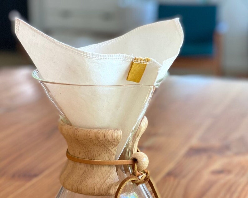 May include: A glass coffee carafe with a light wood collar and a leather tie, featuring a reusable off-white fabric cone filter inserted into the top. The filter has a small, rectangular mustard yellow fabric tag stitched onto its upper edge.