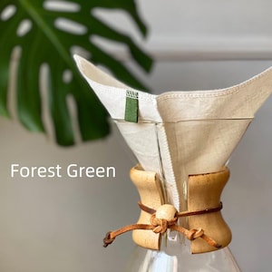 May include: A glass coffee maker with a light wood collar secured by a brown leather cord and a wooden bead. A reusable off-white fabric filter sits inside the glass cone, featuring a small forest green fabric tab. The text Forest Green is visible.