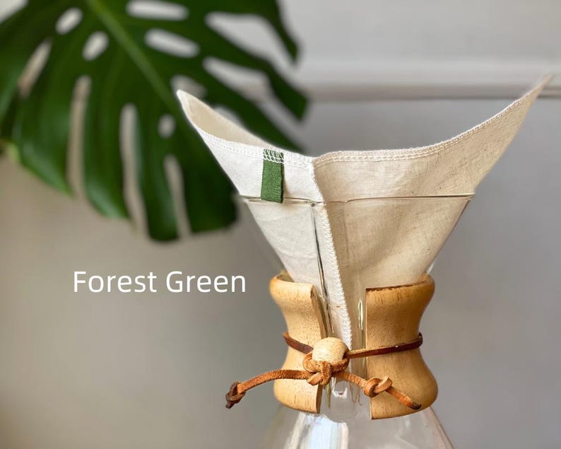 May include: A glass coffee maker with a light wood collar secured by a brown leather cord and a wooden bead. A reusable off-white fabric filter sits inside the glass cone, featuring a small forest green fabric tab. The text Forest Green is visible.