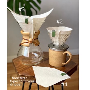 May include: A glass coffee carafe with a wooden collar and leather tie, a ceramic mug with a metal dripper, and three off-white fabric coffee filters with green tabs labeled #2, #3, and #4. The text reads: Three filter types to fit your dripper.
