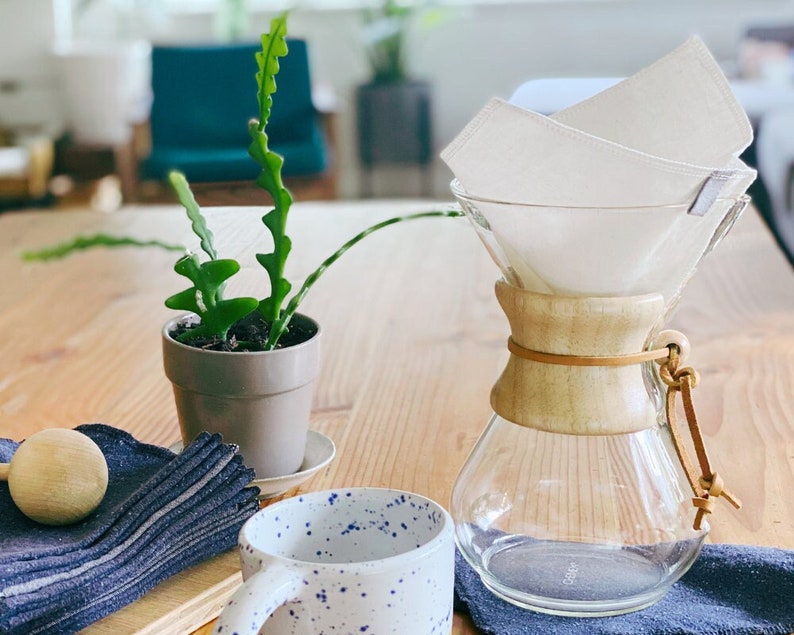 May include: A glass pour-over coffee maker with a light wood collar and a leather tie, featuring a white fabric filter inside. Beside it sits a white ceramic mug with blue speckles, a small potted green plant, and a stack of dark blue fabric.
