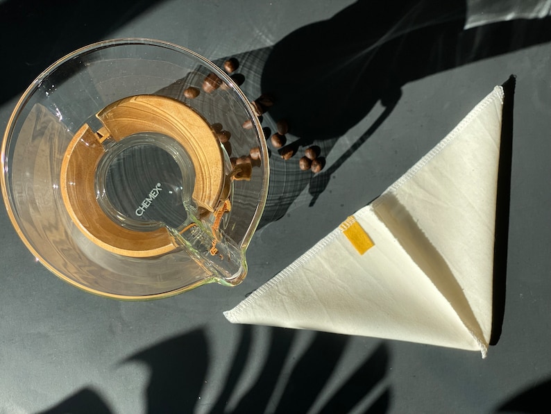 May include: A clear glass Chemex coffee maker with a wooden collar and a small yellow tag, accompanied by a folded off-white fabric cone filter and several roasted coffee beans scattered nearby on a dark surface.