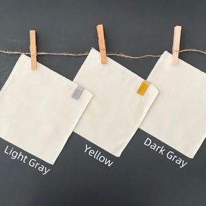 May include: Three square off-white fabric cloths hanging from a twine string with wooden clothespins. Each cloth features a small colored tab on the right edge: light gray, yellow, and dark gray. Text labels below identify the colors.