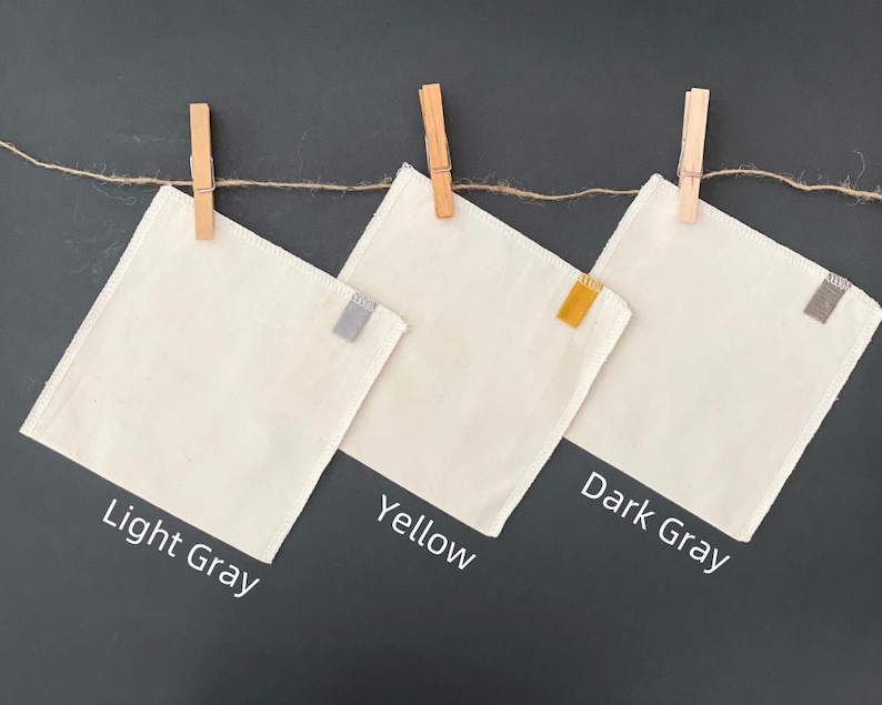 May include: Three square off-white fabric cloths hanging from a twine string with wooden clothespins. Each cloth features a small colored tab on the right edge: light gray, yellow, and dark gray. Text labels below identify the colors.