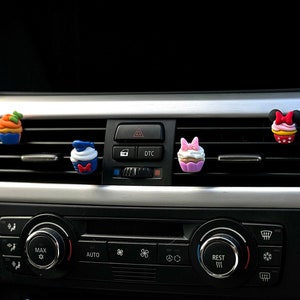 Könnte beinhalten: Vier bunte, cupcakeförmige Autolufterfrischer, die an den Lüftungsschlitzen eines Armaturenbretts hängen. Die Lufterfrischer sind mit Comicfiguren verziert, darunter Mickey Mouse, Minnie Mouse, Donald Duck und ein Kürbis.