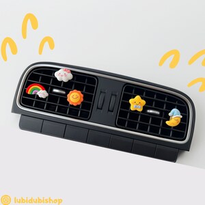 May include: Five colourful car air freshener charms hanging from the air vents of a car. The charms are shaped like a cloud, rainbow, sun, star, and a crescent moon with a sleeping face.