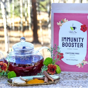 Puede incluir: Una tetera de vidrio llena de té de hierbas rojo, junto a un paquete de té "Immunity Booster". El paquete es rosa con texto blanco y detalles florales. Palitos de canela, cúrcuma y jengibre están en una pequeña bandeja.