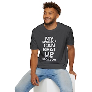 Puede incluir: Camiseta gris oscuro con el texto blanco "MY SPONSOR CAN BEAT UP YOUR SPONSOR". La camiseta la lleva una persona con vaqueros azules. La persona está sentada sobre una superficie blanca.