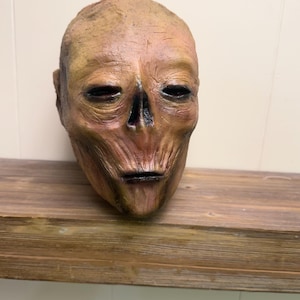 May include: A weathered, human-like mask with a distressed appearance. The mask features closed eyes, a prominent nose, and a slightly open mouth. The color palette includes shades of brown, tan, and black, creating a realistic, aged effect. The mask is resting on a wooden shelf.