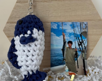 May include: A crocheted blue and white fish keychain with a silver key ring. The fish is standing on a bed of white and gold shredded paper. A photo of a person holding a fish on a boat is in the background.