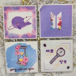 May include: Four square coasters with unique designs. One features a purple whale with a planet, another has a purple logo with cartoon characters, a third shows an hourglass with flowers, and the last has a Korean text and a light stick.
