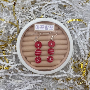 May include: Red and white dangle earrings with a circular design. The earrings are made of three stacked red circles with white stripes. The earrings are displayed on a brown surface inside a white circular container.