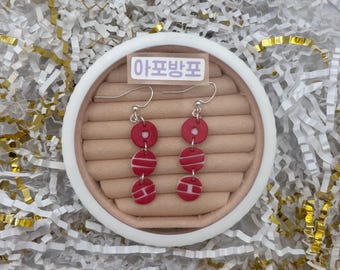 May include: Red and white dangle earrings with a circular design. The earrings are made of three stacked red circles with white stripes. The earrings are displayed on a brown surface inside a white circular container.
