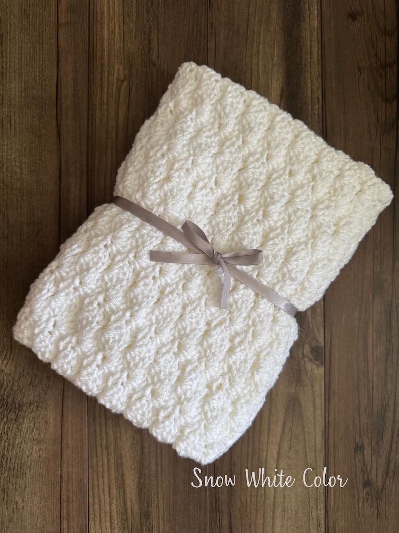 Puede incluir: Una manta de crochet blanca doblada con un patr&oacute;n texturizado, atada con una cinta gris claro. La manta est&aacute; sobre una superficie de madera y el texto "Snow White Color" es visible.