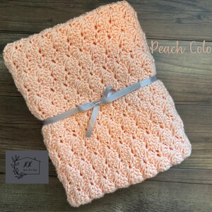 Puede incluir: Una manta de ganchillo color melocot&oacute;n atada con una cinta gris. La manta tiene un patr&oacute;n texturizado y est&aacute; cuidadosamente doblada. Las palabras "Peach Color" est&aacute;n escritas en la esquina superior derecha. Un logotipo est&aacute; en la esquina inferior izquierda.
