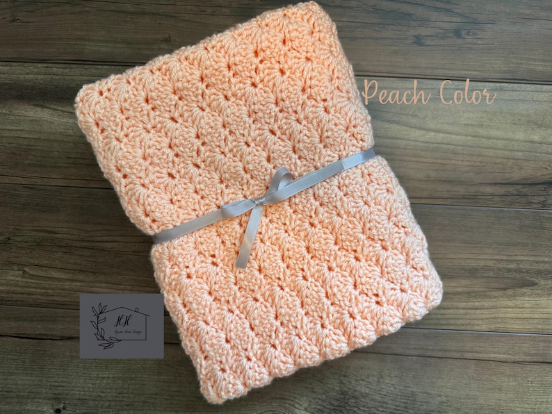 Puede incluir: Una manta de ganchillo color melocot&oacute;n atada con una cinta gris. La manta tiene un patr&oacute;n texturizado y est&aacute; cuidadosamente doblada. Las palabras "Peach Color" est&aacute;n escritas en la esquina superior derecha. Un logotipo est&aacute; en la esquina inferior izquierda.