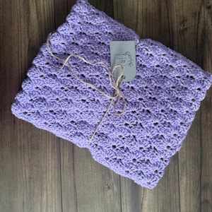 Puede incluir: Una manta de ganchillo plegada, de color lavanda, con un patr&oacute;n de puntada de concha texturizada. Una peque&ntilde;a etiqueta est&aacute; sujeta con hilo. La manta se muestra sobre una superficie de madera oscura.