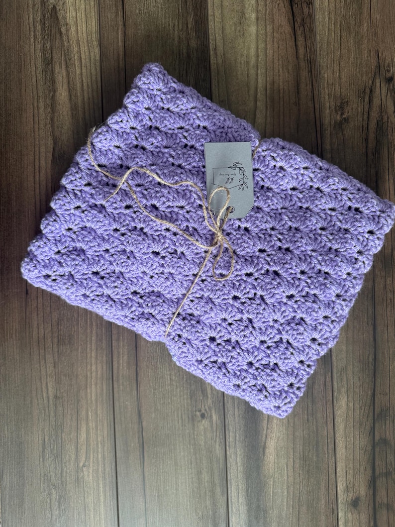 Puede incluir: Una manta de ganchillo plegada, de color lavanda, con un patr&oacute;n de puntada de concha texturizada. Una peque&ntilde;a etiqueta est&aacute; sujeta con hilo. La manta se muestra sobre una superficie de madera oscura.