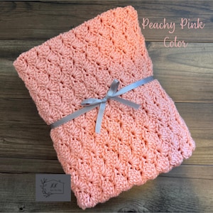 Puede incluir: Una manta de ganchillo rosa melocot&oacute;n atada con una cinta gris. La manta tiene un patr&oacute;n texturizado y las palabras "Peachy Pink Color" est&aacute;n escritas en la parte superior derecha. La manta est&aacute; sobre una superficie de madera.