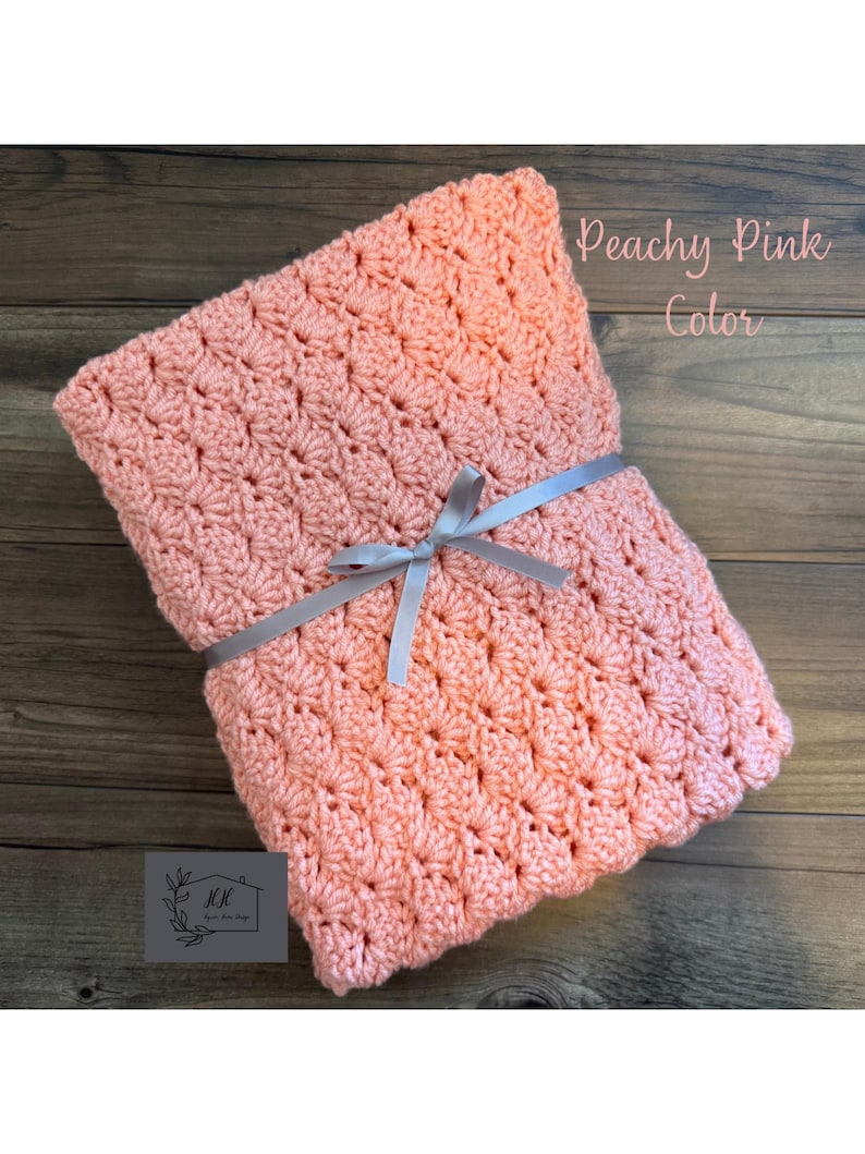Puede incluir: Una manta de ganchillo rosa melocot&oacute;n atada con una cinta gris. La manta tiene un patr&oacute;n texturizado y las palabras "Peachy Pink Color" est&aacute;n escritas en la parte superior derecha. La manta est&aacute; sobre una superficie de madera.