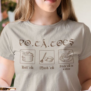 Puede incluir: Una camiseta beige claro con un gráfico marrón que dice "PO.TÄ.TOES" y muestra tres ilustraciones de patatas en preparación. El texto debajo de las ilustraciones dice "Boil 'em, Mash 'em, Stich 'em in a stew."