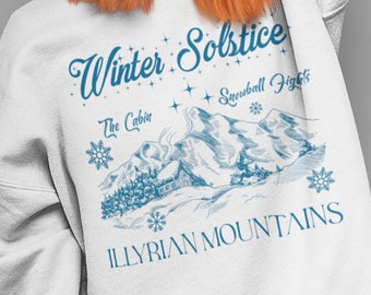May include: White sweatshirt with a blue graphic design featuring a winter scene with mountains, a cabin, snowflakes, and the text "Winter Solstice The Cabin Snowball Fights Illyrian Mountains".