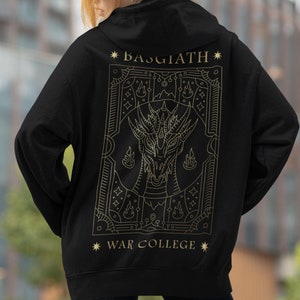 Peut inclure: Sweat-shirt à capuche noir avec un motif doré d'un dragon dans un cadre avec le texte "BASGIATH" et "WAR COLLEGE" au dos.