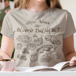 Puede incluir: Una camiseta gris claro con un gráfico marrón de alimentos y el texto "What about Second Breakfast?" impreso en la parte delantera.