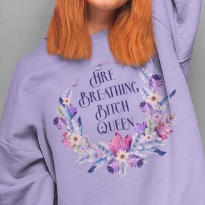 Op de afbeelding: Een lila sweatshirt met de tekst "FIRE BREATHING BITCH QUEEN" in een cirkelvormig ontwerp. Het ontwerp bevat bloemen, veren en kristallen in tinten paars, roze en blauw.