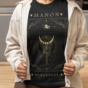 May include: Black t-shirt with a gold graphic design featuring a crescent moon, stars, and the text "*MANON*" and "*BLACKBEAK*"