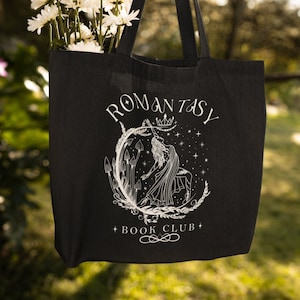May include: Black tote bag featuring the text "ROMANTASY BOOK CLUB" and a white illustration of a woman with a crown, surrounded by mushrooms and stars. The bag is filled with white flowers and hangs from a tree branch.