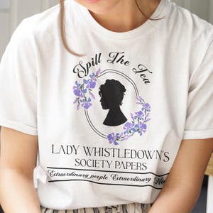 Op de afbeelding: Wit T-shirt met de tekst "Spill The Tea" en "Lady Whistledown's Society Papers". Het ontwerp bevat een silhouet en bloemaccenten. De persoon draagt een bril en zit op een stoel.
