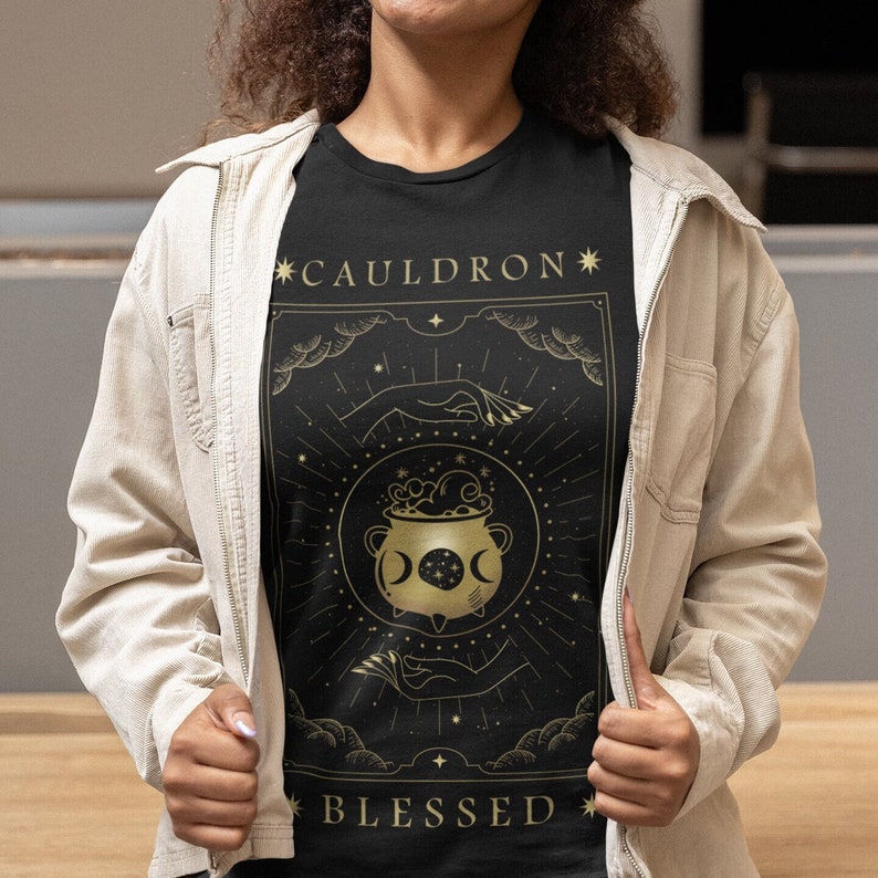 May include: Black t-shirt with a gold graphic of a cauldron with a crescent moon and stars. The text "*CAULDRON*" and "*BLESSED*" is printed in gold on the shirt.