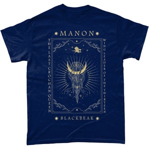 May include: A navy blue t-shirt with a gold design featuring a crescent moon, stars, and a dragon. The text "*MANON* THE LAST CROCHAN QUEEN" and "*BLACKBEAK* WING LEADER OF THE THIRTEEN" is printed on the shirt.