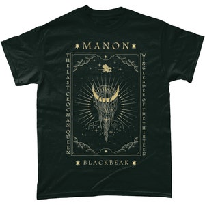 May include: Black t-shirt with a gold graphic design featuring a crescent moon, stars, and the text "*MANON* THE LAST CROCHAN QUEEN WING LEADER OF THE THIRTEEN *BLACKBEAK*"