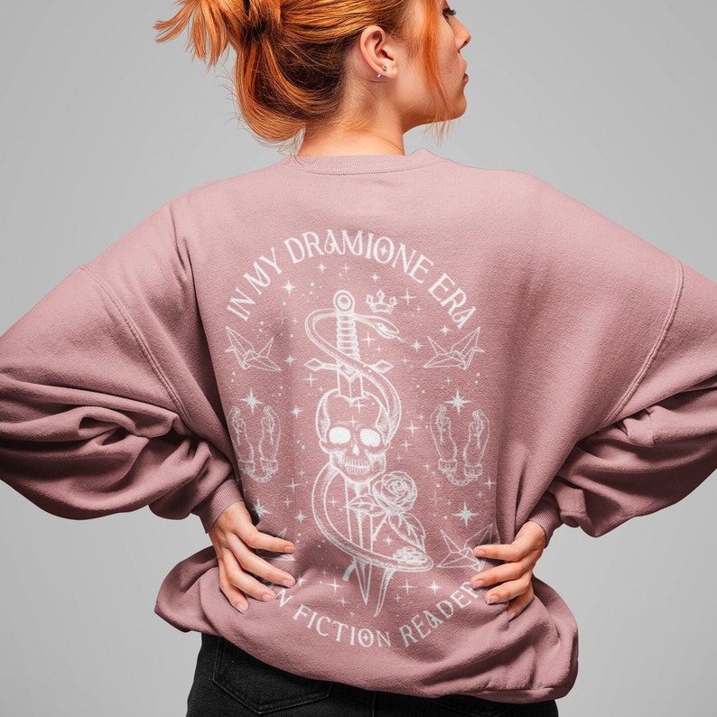 K&ouml;nnte beinhalten: Ein rosafarbenes Sweatshirt mit einem wei&szlig;en Grafikdesign auf der R&uuml;ckseite. Das Design zeigt einen Sch&auml;del mit einem Schwert und einer Schlange, umgeben von Sternen und Origami-Kranichen. Der Text "In My Dramione Era" und "Fiction Reader" ist ebenfalls im Design enthalten.
