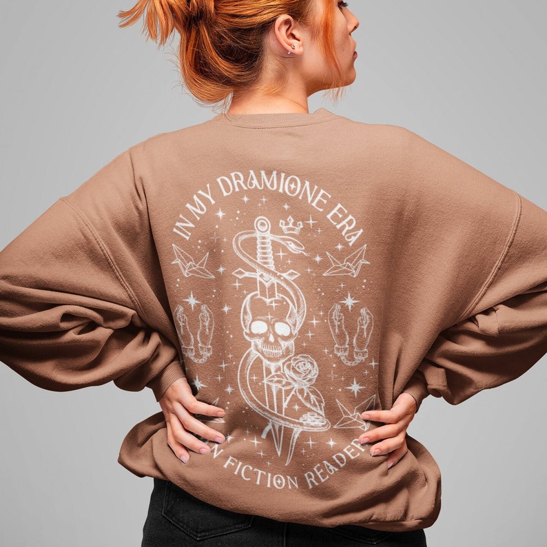 K&ouml;nnte beinhalten: Ein braunes Sweatshirt mit einem wei&szlig;en Grafikdruck eines Sch&auml;dels mit einer Schlange, die sich um ein Schwert windet. Der Text "In My Dramione Era" befindet sich &uuml;ber dem Grafikdruck und "A Non Fiction Reader" darunter.